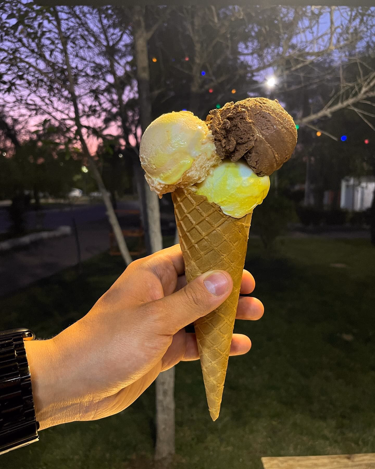 Cono de barquillo con tres bolas de helado sostenido por una mano, con árboles y luces de colores detrás