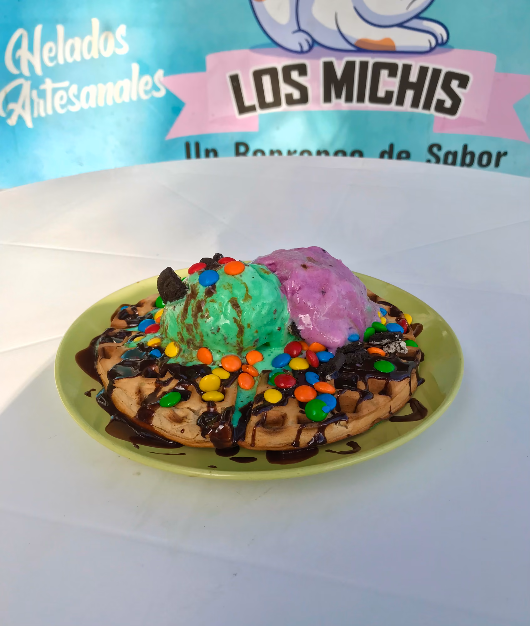 Waffle con chocolate, bolitas de colores y dos bolas de helado, frente al letrero de Los Michis